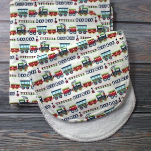May include: A baby bib and matching receiving blanket with a repeating pattern of colorful toy trains on a white background. The text "CHOO CHOO" is printed throughout the design. The bib has a white terrycloth backing.