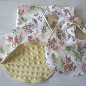 May include: A set of baby items featuring a woodland animal print. The set includes a blanket, a burp cloth, and a bib. The blanket is yellow on the back and has a white and green woodland animal print on the front. The burp cloth and bib are white with a green and white woodland animal print.