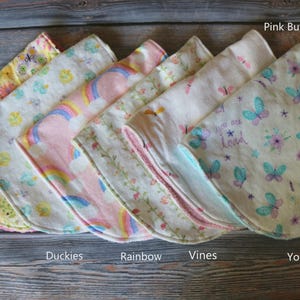 May include: A stack of seven baby burp cloths with various patterns. Designs include yellow floral, duckies, rainbows, vines, and pink butterflies. One cloth has the text "You are Loved". The cloths are made of soft fabric.
