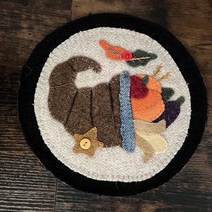 May include: A round wool rug with a black border features a brown cornucopia filled with colorful fruits and vegetables. The cornucopia is surrounded by a white background with a small yellow star with a button in the center.