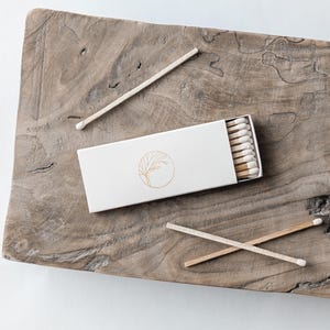 May include: A white matchbox with a gold tree logo, partially open to reveal the match heads. Two matches are crossed, and one is placed separately on a rustic wooden surface. The matches have white tips.
