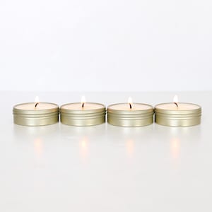 May include: Four round, gold-colored metal tins containing lit candles arranged in a horizontal row. Each candle features a visible flame with a glowing wick, set within a shallow, circular container with a smooth, metallic finish and a simple lid rim.