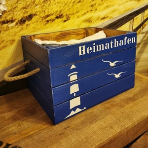 Könnte beinhalten: Eine blaue Holzkiste mit einem Seilgriff, bemalt mit einem weißen Leuchtturm und Möwen. Die Kiste trägt den Schriftzug "Heimathafen" auf der Vorderseite.