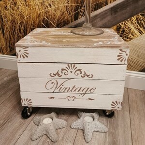 Könnte beinhalten: Weiße Holzkiste mit Rollen, verziert mit braunen Details und dem Wort "Vintage". Die Kiste steht auf einem Holzboden mit zwei Kerzenhaltern in Form von Seesternen.