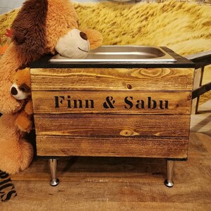 Könnte beinhalten: Ein Futter- und Wassernapfständer aus Holz mit einer Schüssel aus Edelstahl. Der Ständer hat eine rustikale braune Oberfläche und ist mit dem Text "Finn & Sabu" verziert.