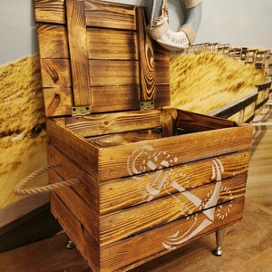 May include: A wooden storage box with a rope handle and metal legs. The box has a distressed wood finish and a white anchor design. The lid is open and a life preserver with the text "Welcome On Board" is hanging above the box.
