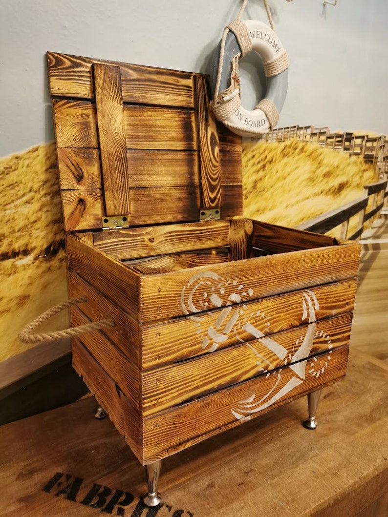 May include: A wooden storage box with a rope handle and metal legs. The box has a distressed wood finish and a white anchor design. The lid is open and a life preserver with the text "Welcome On Board" is hanging above the box.
