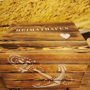 May include: A wooden storage box with a white painted anchor and heart design. The box has the words "HEIMATHAFEN" painted on the top.