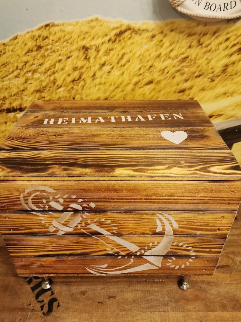 May include: A wooden storage box with a white painted anchor and heart design. The box has the words "HEIMATHAFEN" painted on the top.