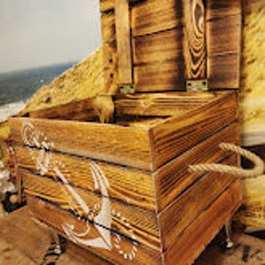 May include: A wooden storage box with a rope handle and an anchor design. The box is painted in a light brown color and has a distressed finish. The box is sitting on a wooden surface.