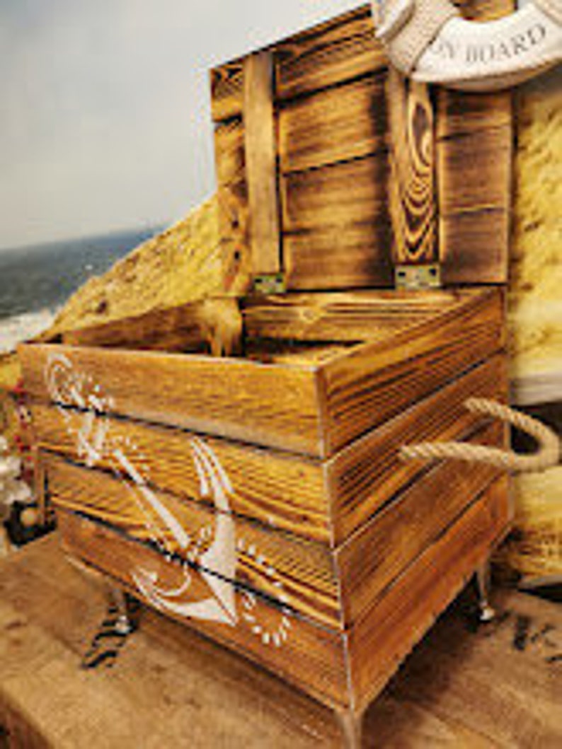 May include: A wooden storage box with a rope handle and an anchor design. The box is painted in a light brown color and has a distressed finish. The box is sitting on a wooden surface.
