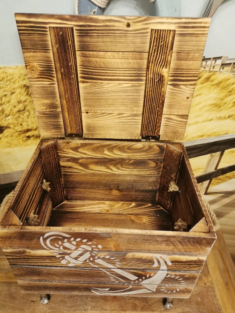 May include: A wooden storage box with a hinged lid and rope handles. The box is decorated with a white anchor design and has a distressed wood finish.