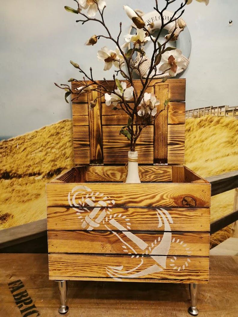 May include: A wooden storage box with a hinged lid and metal legs. The box is decorated with a white anchor design and has a distressed wood finish.