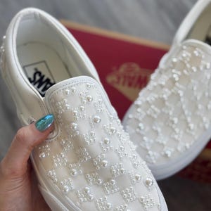 May include: White slip-on Vans sneakers with a pearl design. The pearls are arranged in a checkered pattern.