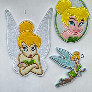 Może przedstawiać: Trzy haftowane łatki do prasowania z Tinkerbell, wróżką z Disneya. Pierwsza łatka przedstawia Tinkerbell z założonymi rękami, druga pokazuje jej głowę i ramiona, a trzecia przedstawia ją w locie z rozłożonymi skrzydłami.