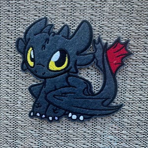 Könnte beinhalten: Ein schwarzer Drachenaufnäher mit gelben Augen und einer roten Schwanzflosse. Der Drache sitzt mit leicht ausgebreiteten Flügeln. Der Aufnäher ist aus Filz und hat weiße Akzente an den Füßen.