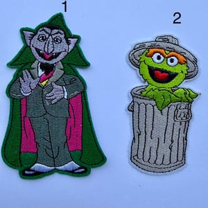 May include: Two embroidered patches labelled 1 and 2. Patch 1 depicts a vampire character in a grey suit and green cape with a pink lining. Patch 2 depicts a green furry character with orange eyebrows inside a grey trash can with a lid.