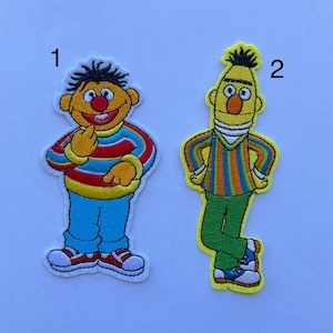 May include: Two embroidered patches featuring cartoon characters. One patch depicts a character in a striped shirt, blue pants, and red shoes. The other patch shows a character in a striped shirt, green pants, and blue shoes. Both patches have yellow borders.