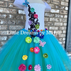 Puede incluir: Un vestido de tul de color turquesa con adornos de flores de colores y un corpiño blanco con encaje negro y un detalle de mariposa. El vestido está en un maniquí blanco. "Little Dreams by Mayra" está escrito en el vestido.