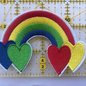 Puede incluir: Parche bordado con un arcoíris con arcos rojos, naranjas, amarillos, verdes y azules. Debajo del arcoíris hay cuatro diseños en forma de corazón en azul, verde, rojo y amarillo. El parche tiene un borde blanco.