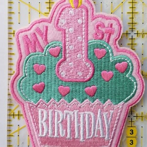 Puede incluir: Cupcake de fieltro rosa con glaseado blanco y una vela rosa con llama amarilla. El cupcake tiene glaseado verde con corazones rosas. El texto "My 1st Birthday" está bordado en blanco sobre el glaseado.