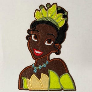 May include: Embroidered patch featuring a cartoon princess with brown skin, wearing a green and yellow crown and dress. The princess has a red smile and a blue necklace. The patch is designed with a felt-like texture.