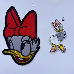 May include: Two embroidered iron-on patches featuring a cartoon duck with a red bow and a cartoon duck with a pink bow. The patches are on a white background.
