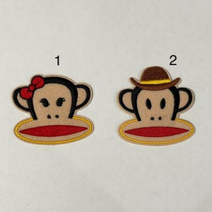 May include: Two embroidered monkey face patches. One has a red bow, the other a brown hat. Both have tan faces, black outlines, red mouths, and yellow and red accents. The patches are numbered 1 and 2.