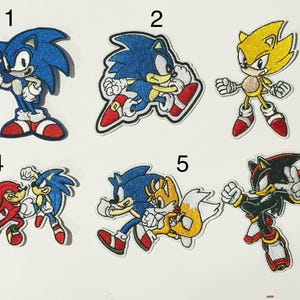 Pode incluir: Seis emblemas bordados com personagens de Sonic the Hedgehog. O número 1 mostra o Sonic azul em pé. O 2 mostra o Sonic azul correndo. O 3 mostra o Super Sonic amarelo. O 4 mostra Knuckles e Sonic. O 5 mostra Sonic e Tails. O 6 mostra Shadow.