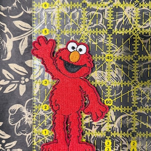 May include: A red Elmo patch with white eyes, a black mouth, and an orange nose. The patch is approximately 3 inches tall. The patch is on a yellow grid with a floral background.