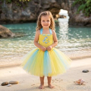May include: A yellow and light blue tutu dress featuring a crocheted bodice with a small embroidered fish character on the chest and a multi-layered tulle skirt in alternating yellow and light blue colors with light blue halter straps.