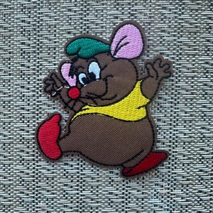 May include: An embroidered patch of a brown mouse wearing a green hat and a yellow shirt. The mouse is walking and waving with its right hand.