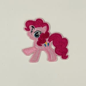 May include: An embroidered iron-on patch featuring a light pink pony character with a bright magenta curly mane and tail. The character has large blue eyes, a smiling expression, and a small blue and yellow balloon symbol on its hind leg.