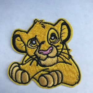 May include: Embroidered patch featuring a yellow lion cub with black outlines and a pink nose. The lion cub is in a seated position with its paws in front. The patch is likely for clothing or accessories.