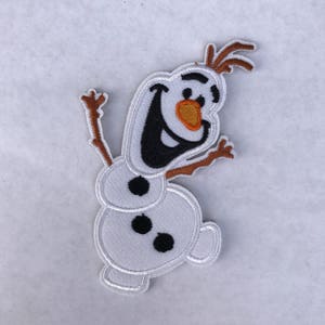 May include: A white felt applique of Olaf, a snowman character from the movie Frozen, with a brown felt stick figure body and orange felt nose. He is smiling with black felt eyes and mouth.