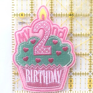 May include: A pink and green embroidered patch in the shape of a cupcake. The patch says "MY 2nd BIRTHDAY" with a candle on top. The cupcake has pink hearts and a pink outline.