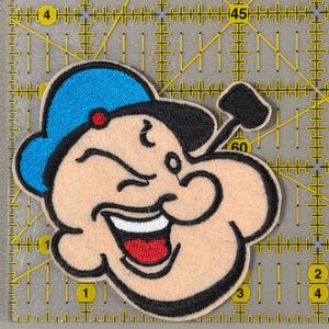 May include: Embroidered patch featuring a smiling cartoon character with a blue hat, black hair, and a red mouth. The patch has a beige background and black outline. The character is holding a black hammer.