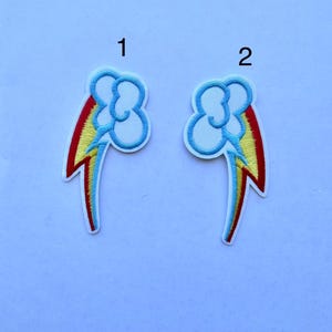 Op de afbeelding: Twee geborduurde patches met een blauwe wolk, regenboog en bliksemontwerp. De patches hebben een witte rand en zijn genummerd 1 en 2. De regenboog heeft de kleuren rood, geel en blauw.