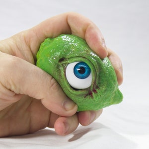 May include: A green, textured, squishy toy with a large blue eye. The toy is being held in a hand.