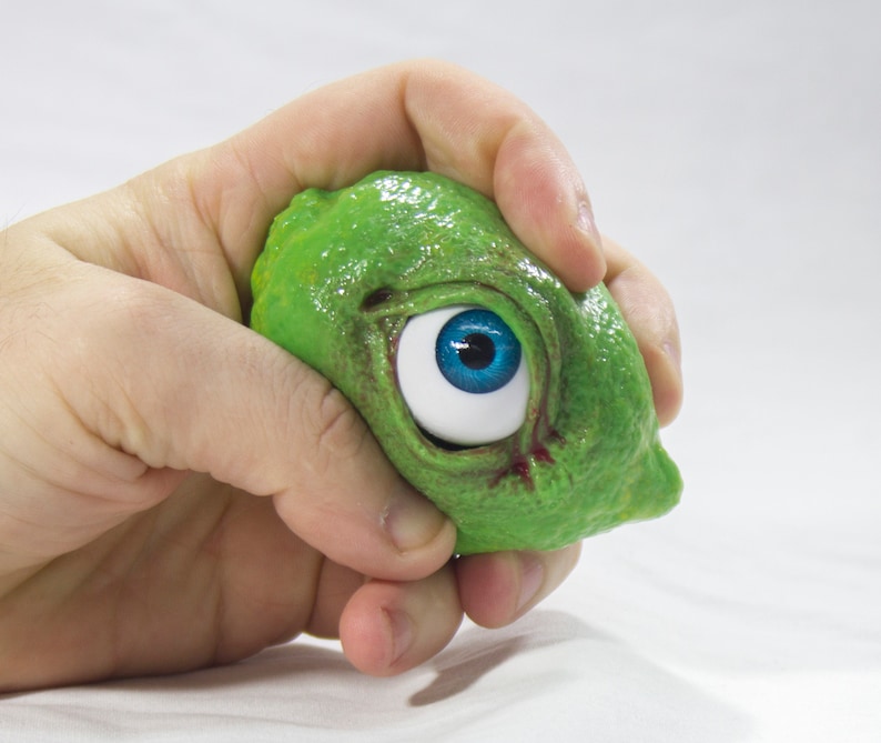 May include: A green, textured, squishy toy with a large blue eye. The toy is being held in a hand.