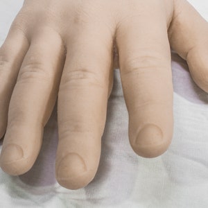 May include: A close-up of a pale, flesh-toned hand with five fingers. The hand is lying flat on a white surface. The fingers are slightly curled, and the nails are trimmed and rounded.