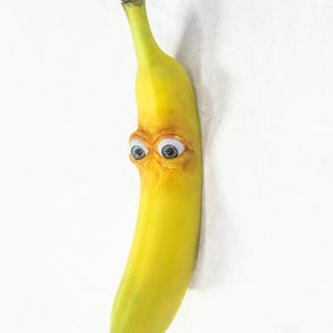 May include: A quirky, yellow banana with a face, complete with two blue eyes and a surprised expression. The banana has a green stem and a brown tip, set against a white backdrop.