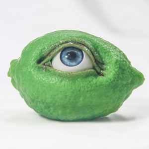 Puede incluir: Un limón verde con un ojo azul pintado en él. El ojo está abierto y mirando hacia la derecha.