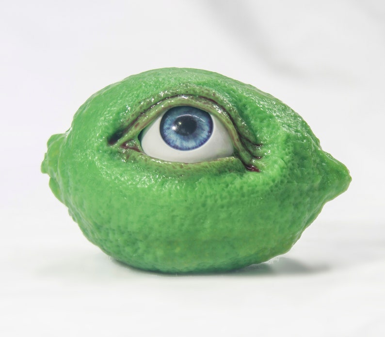 May include: A green lime with a blue eye painted on it. The eye is open and looking to the right.