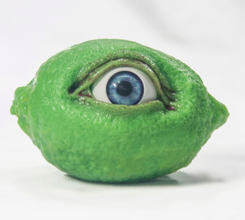 May include: A green lime with a blue eye embedded in it. The eye is open and looking to the right.