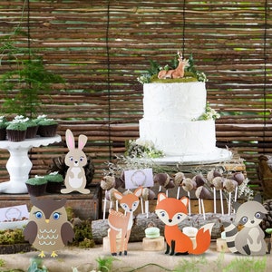 May include: A woodland-themed party table setting with a white two-tier cake decorated with a deer figurine. The table is decorated with cupcakes, cookies, and various animal figurines, including a fox, owl, deer, rabbit, and raccoon.