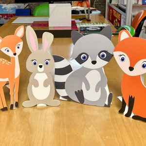 May include: A set of six woodland animal cutouts, including an owl, deer, rabbit, raccoon, and fox. The cutouts are made of cardboard and are painted in various shades of brown, gray, and orange. The animals have large, cartoonish eyes and are standing on a wooden table.