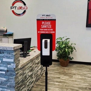Könnte beinhalten: Ein schwarzer Händedesinfektionsmittelspender mit einem roten Schild mit der Aufschrift "BITTE DESINFIZIEREN SIE IHRE HÄNDE, BEVOR SIE ZUR ARBEIT ZURÜCKKEHREN". Der Spender steht auf einem schwarzen Ständer. Das Bild zeigt auch einen Empfangstresen und ein gerahmtes Trikot.