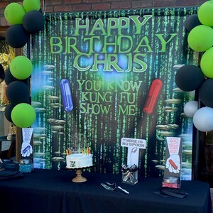 May include: A green and black birthday banner with the text "Happy Birthday Chris" and "You Know Kung Fu! Show Me!"  The banner is decorated with green and black balloons and a cake with candles. There are also two signs that say "There is no spoon" and "Red Pill Blue Pill".