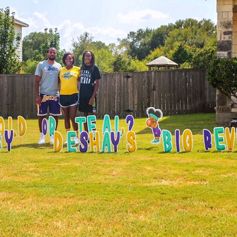 May include: A family of three stands in front of a gender reveal party sign that reads "Boy or Teal? Deshay's Big Reveal". The sign is made of large, colorful letters and features a cartoon baby holding a basketball. The family is dressed in yellow and purple Lakers jerseys.
